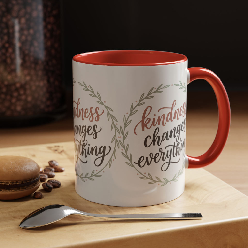 Inspirational ceramic accent coffee mug featuring hand-lettered “Kindness Changes Everything” typography surrounded by a delicate laurel wreath in soft sage and blush tones.