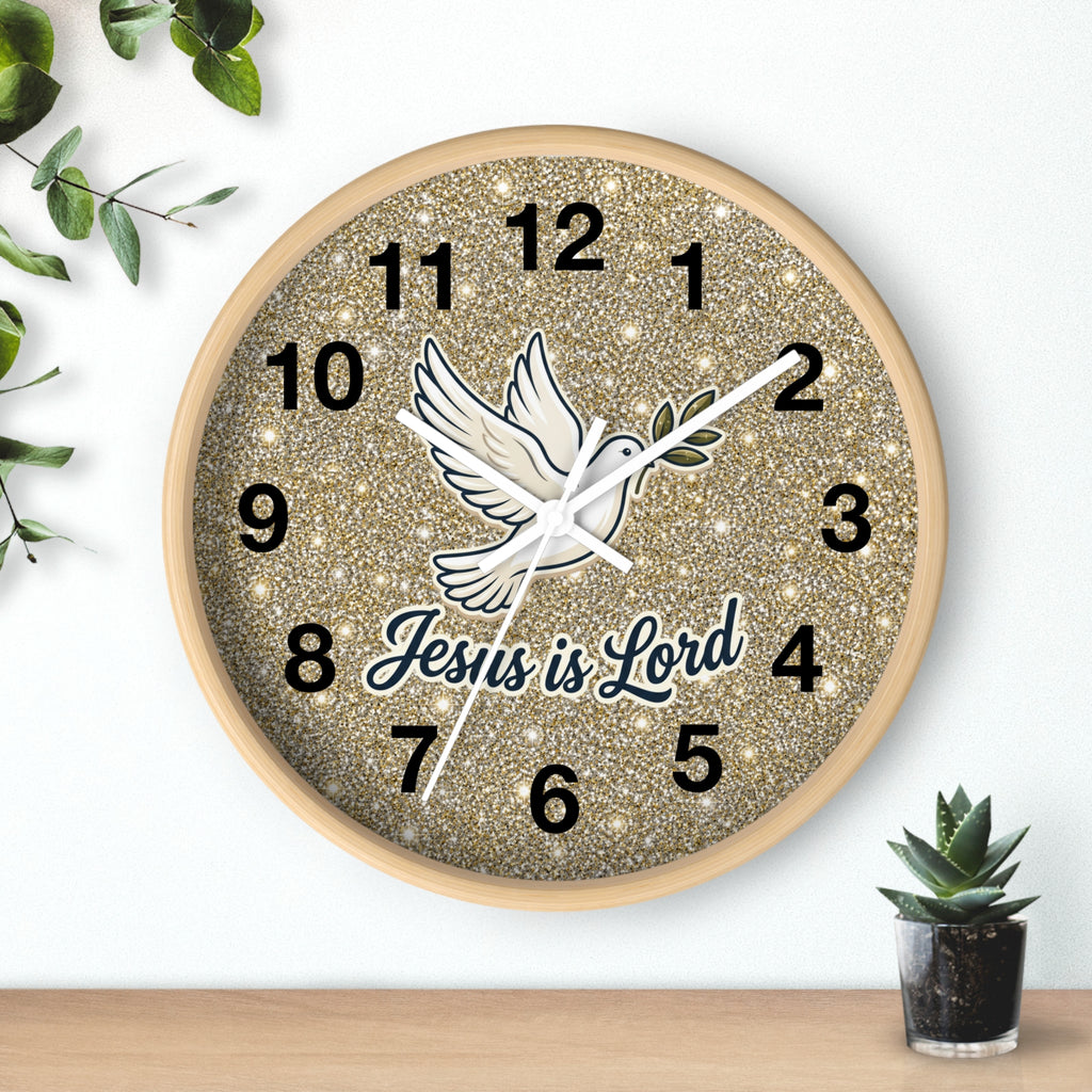 A round wall clock with a glittery gold face, black Arabic numerals, a white dove illustration, and the text 'Jesus is Lord' in dark blue script. The clock is mounted on a white wall above a light wood surface, with small green plants in the corners.