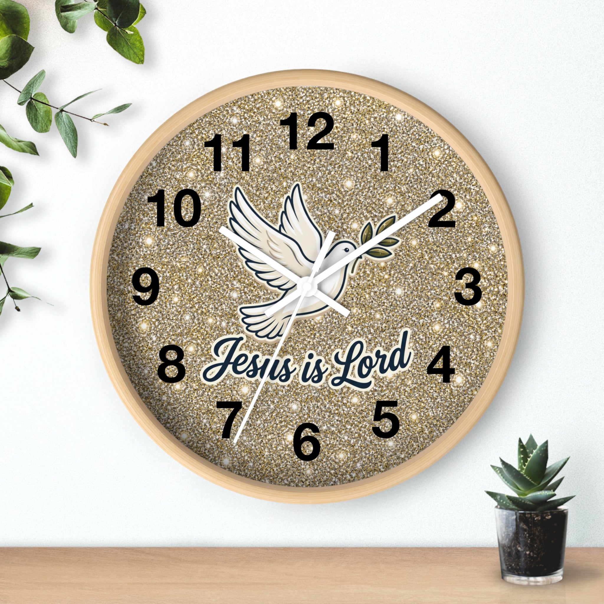 A round wall clock with a glittery gold face, black Arabic numerals, a white dove illustration, and the text 'Jesus is Lord' in dark blue script. The clock is mounted on a white wall above a light wood surface, with small green plants in the corners.