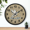 A round wall clock with a black frame and a glittery gold face, featuring a white dove illustration and the text 'Jesus is Lord' in blue and black script. The clock is shown hanging on a white wall above a wooden surface with small green plants nearby.