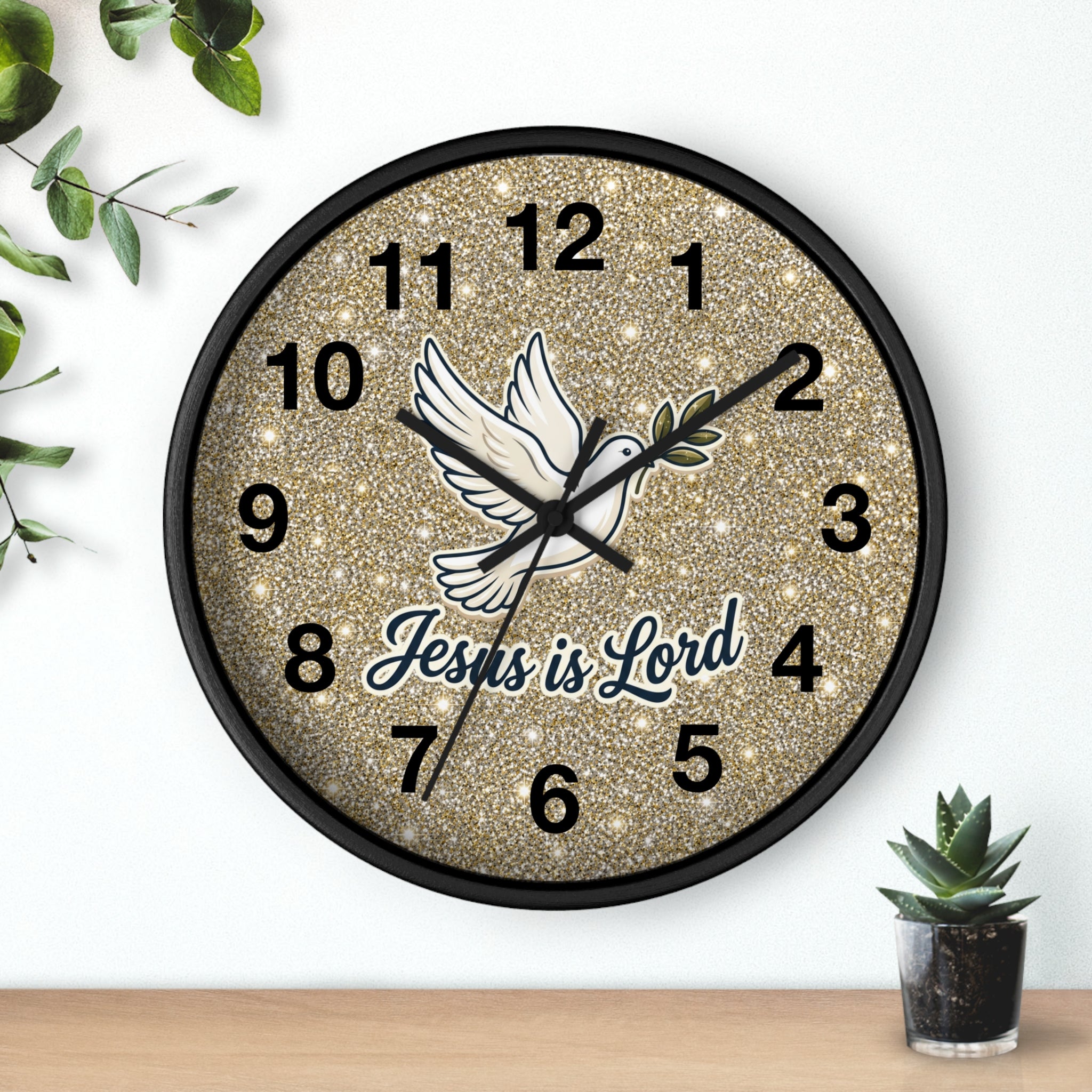 A round wall clock with a black frame and a glittery gold face, featuring a white dove illustration and the text 'Jesus is Lord' in blue and black script. The clock is shown hanging on a white wall above a wooden surface with small green plants nearby.