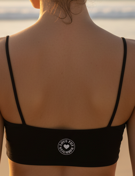 Women's luxe athletic crop bra featuring a "Love Thy Neighbor" heart emblem with double spaghetti straps and medium-support recycled performance fabric.