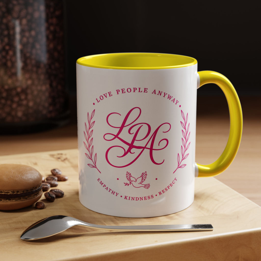 Inspirational ceramic coffee mug featuring soft pink “Love People Anyway” lettering, laurel accents, a dove motif, and contrasting yellow handle and interior.