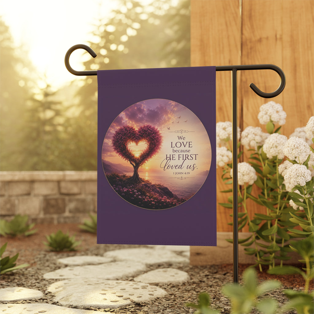 Inspirational Christian garden flag featuring a heart-shaped tree at sunset with the scripture “We Love Because He First Loved Us” in warm glowing tones.