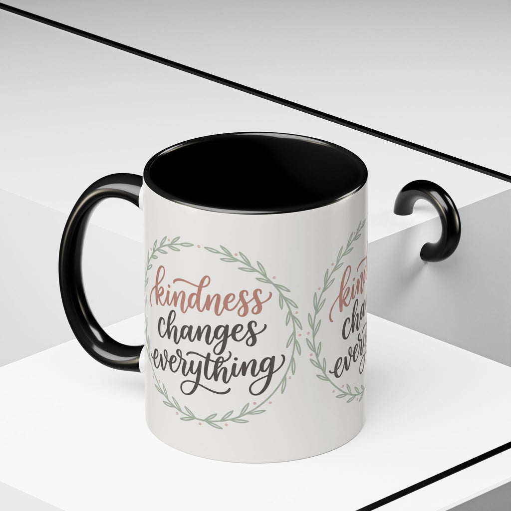 Inspirational ceramic accent coffee mug featuring hand-lettered “Kindness Changes Everything” typography surrounded by a delicate laurel wreath in soft sage and blush tones.