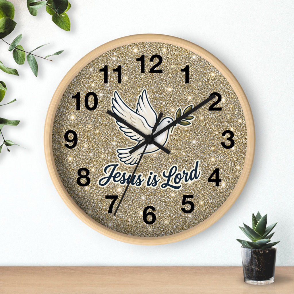 A round wall clock with a glittery gold face, black Arabic numerals, a white dove illustration, and the words 'Jesus is Lord' in script. The clock is mounted on a white wall above a light wood surface, with small green plants in the corners.