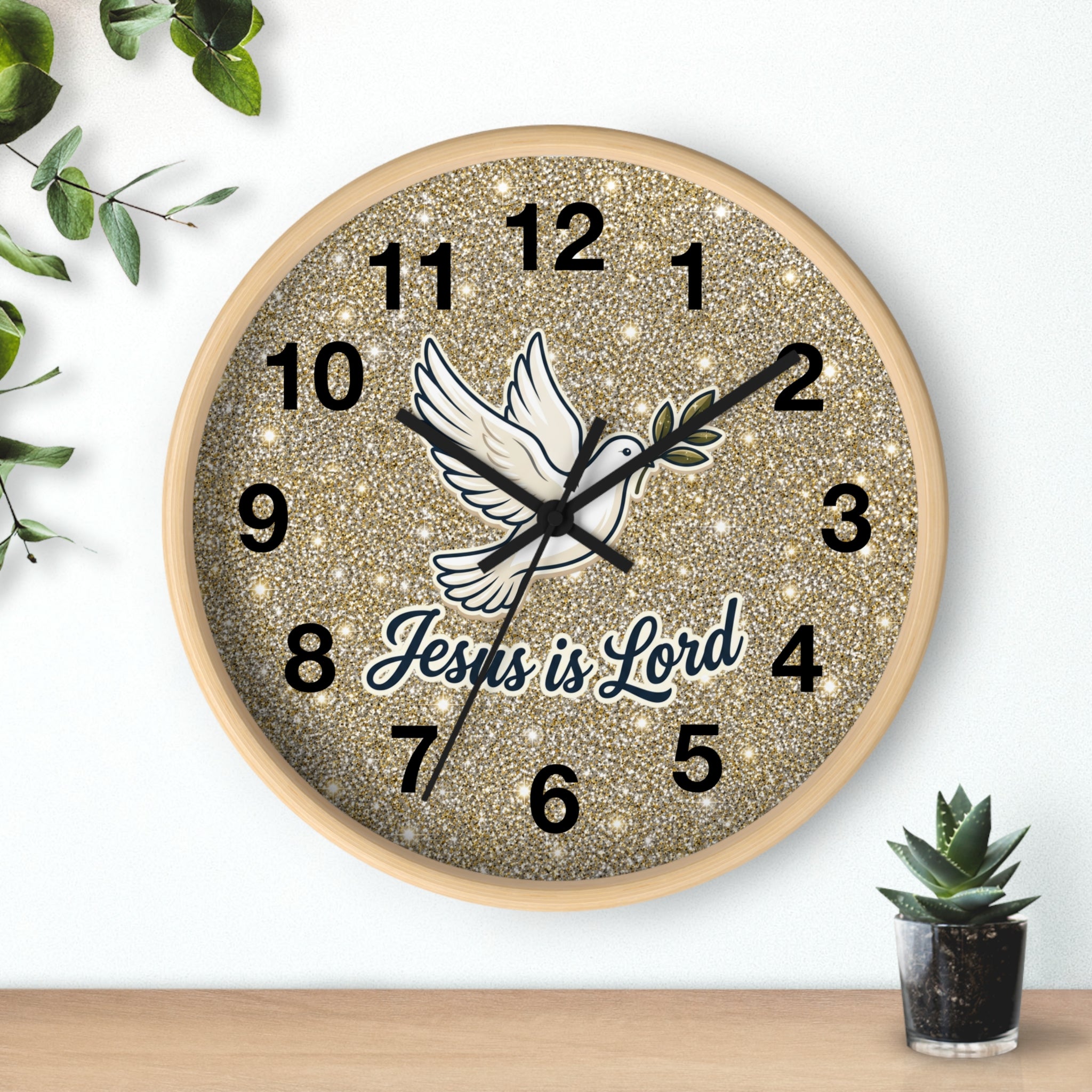 A round wall clock with a glittery gold face, black Arabic numerals, a white dove illustration, and the words 'Jesus is Lord' in script. The clock is mounted on a white wall above a light wood surface, with small green plants in the corners.
