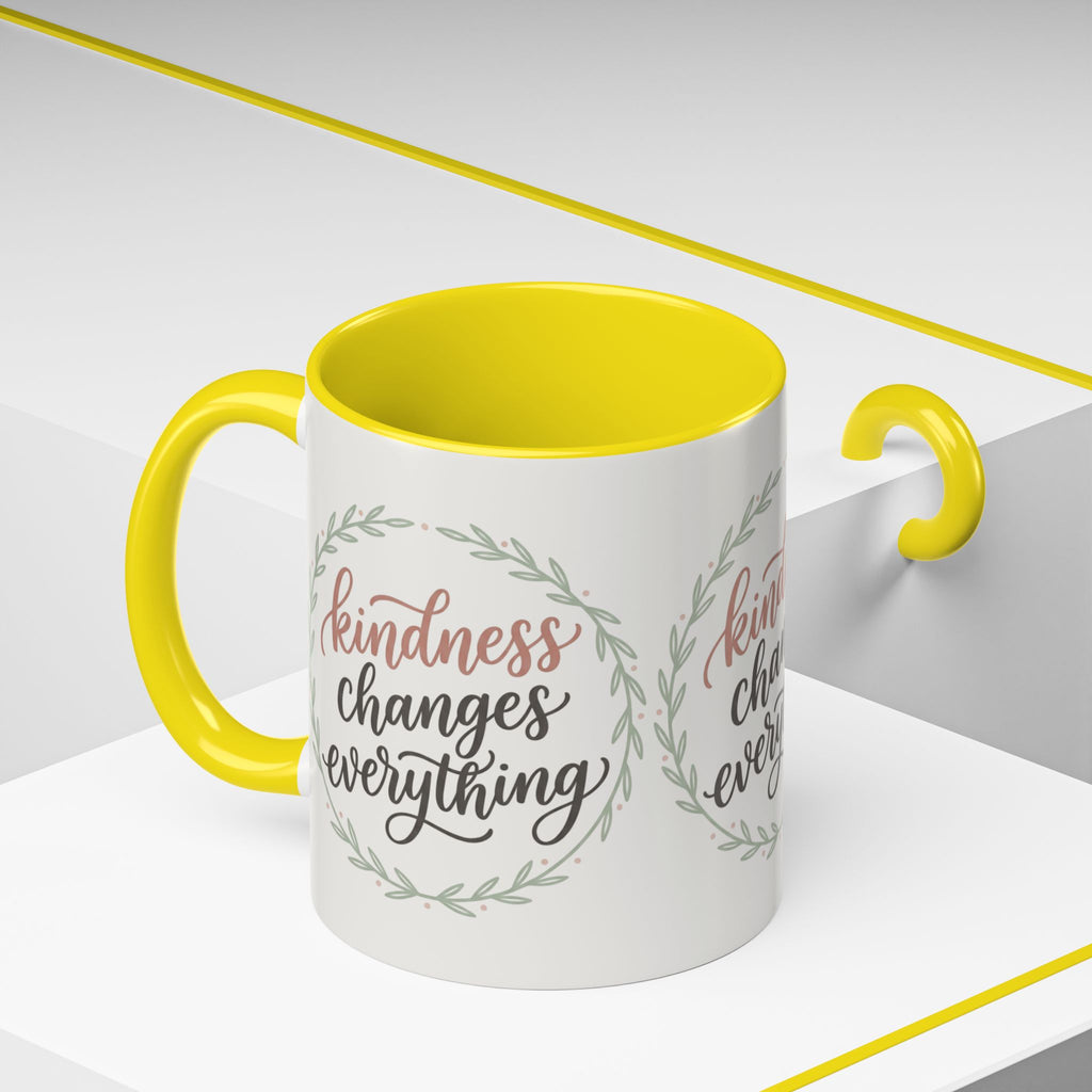 Inspirational ceramic accent coffee mug featuring hand-lettered “Kindness Changes Everything” typography surrounded by a delicate laurel wreath in soft sage and blush tones.
