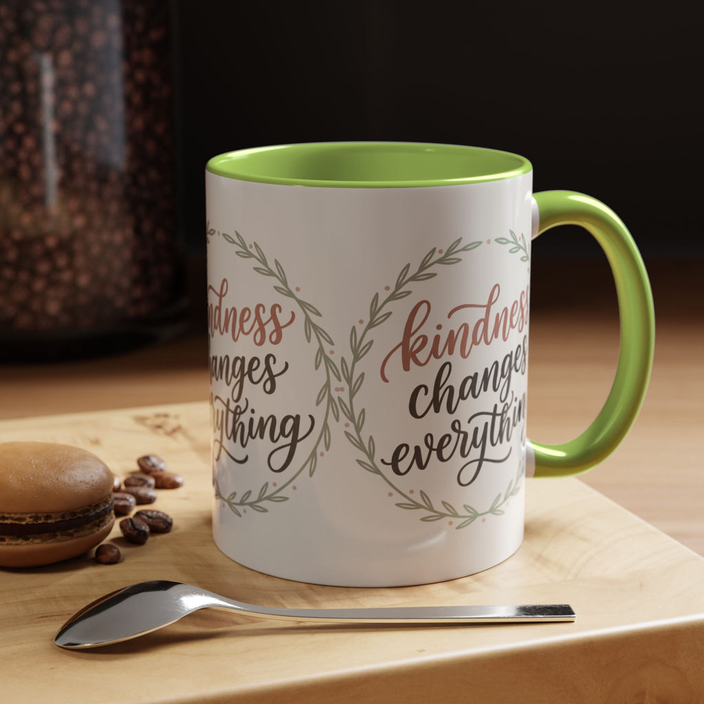 Inspirational ceramic accent coffee mug featuring hand-lettered “Kindness Changes Everything” typography surrounded by a delicate laurel wreath in soft sage and blush tones.