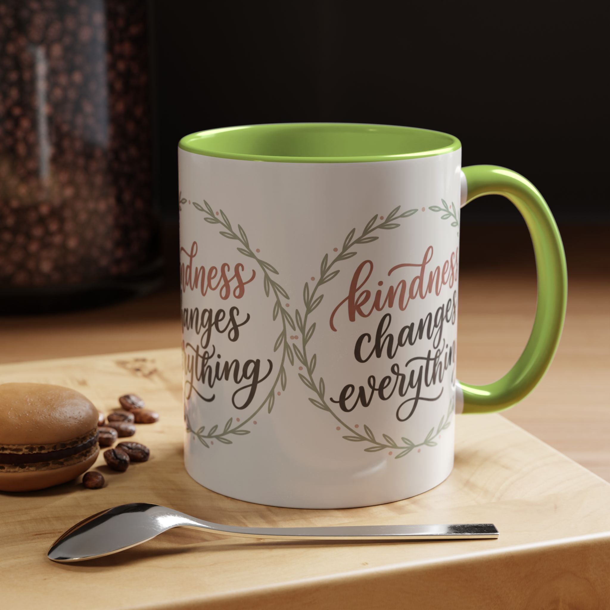 Inspirational ceramic accent coffee mug featuring hand-lettered “Kindness Changes Everything” typography surrounded by a delicate laurel wreath in soft sage and blush tones.