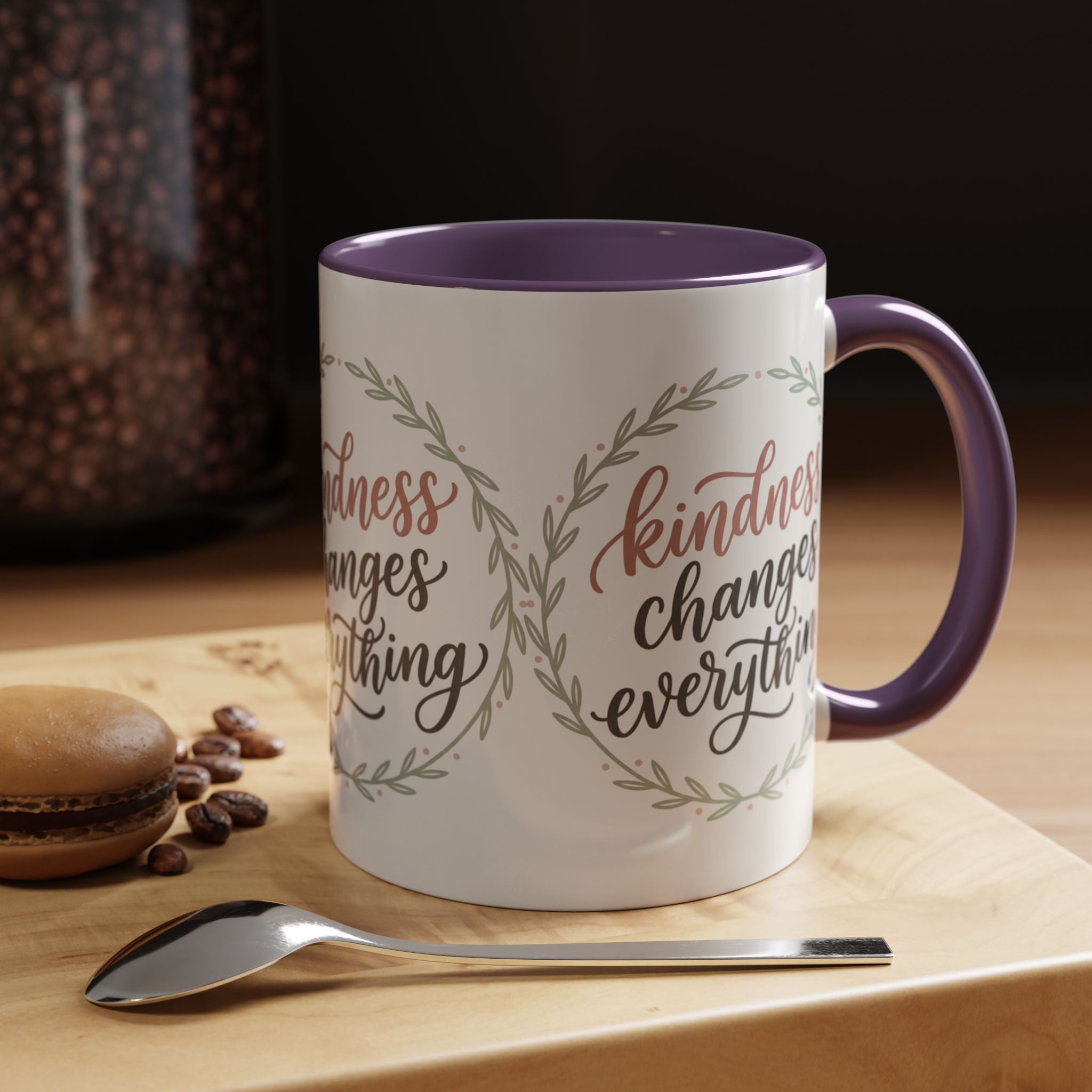 Inspirational ceramic accent coffee mug featuring hand-lettered “Kindness Changes Everything” typography surrounded by a delicate laurel wreath in soft sage and blush tones.