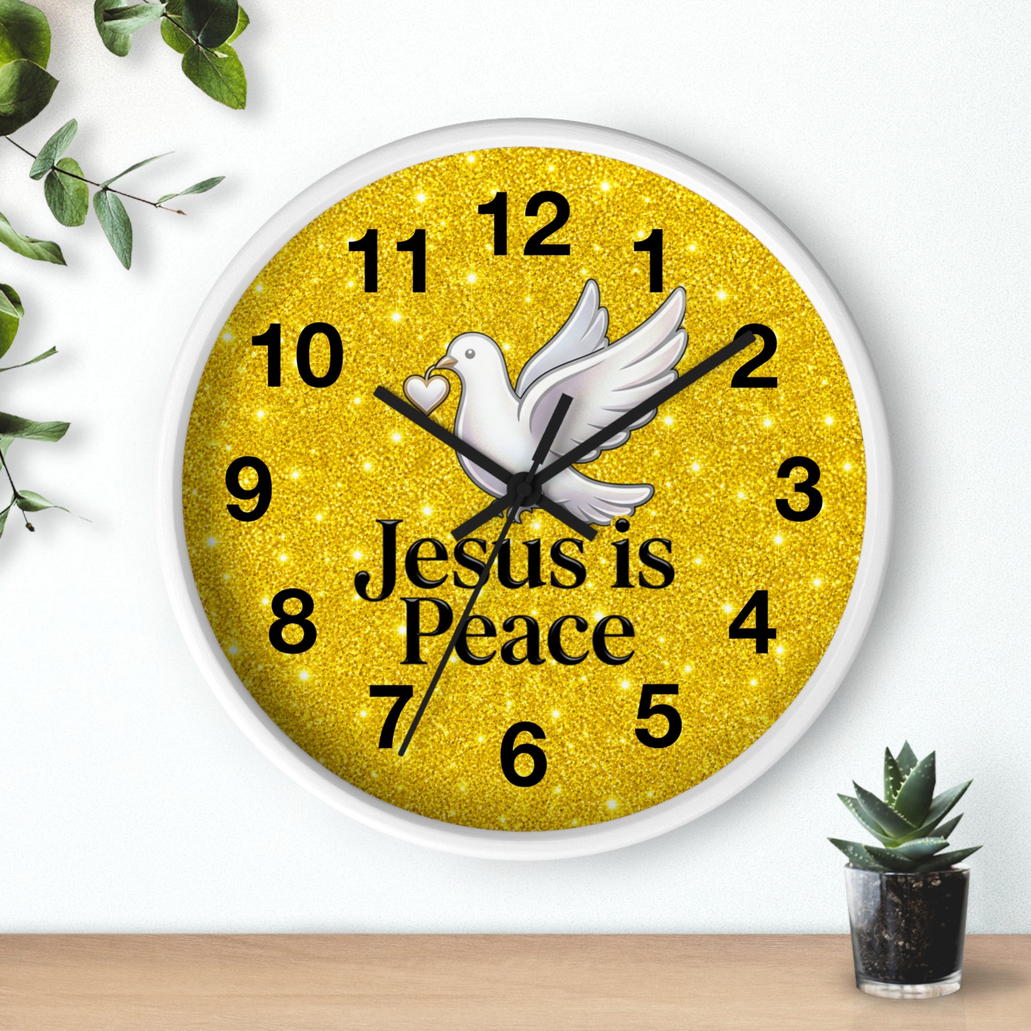 A round wall clock with a white frame and a glittery gold face, featuring a white dove, black numbers, and the words 'Jesus is Peace' in black text. The clock is mounted on a white wall above a light wood surface, with decorative green leaves and a small succulent plant nearby.