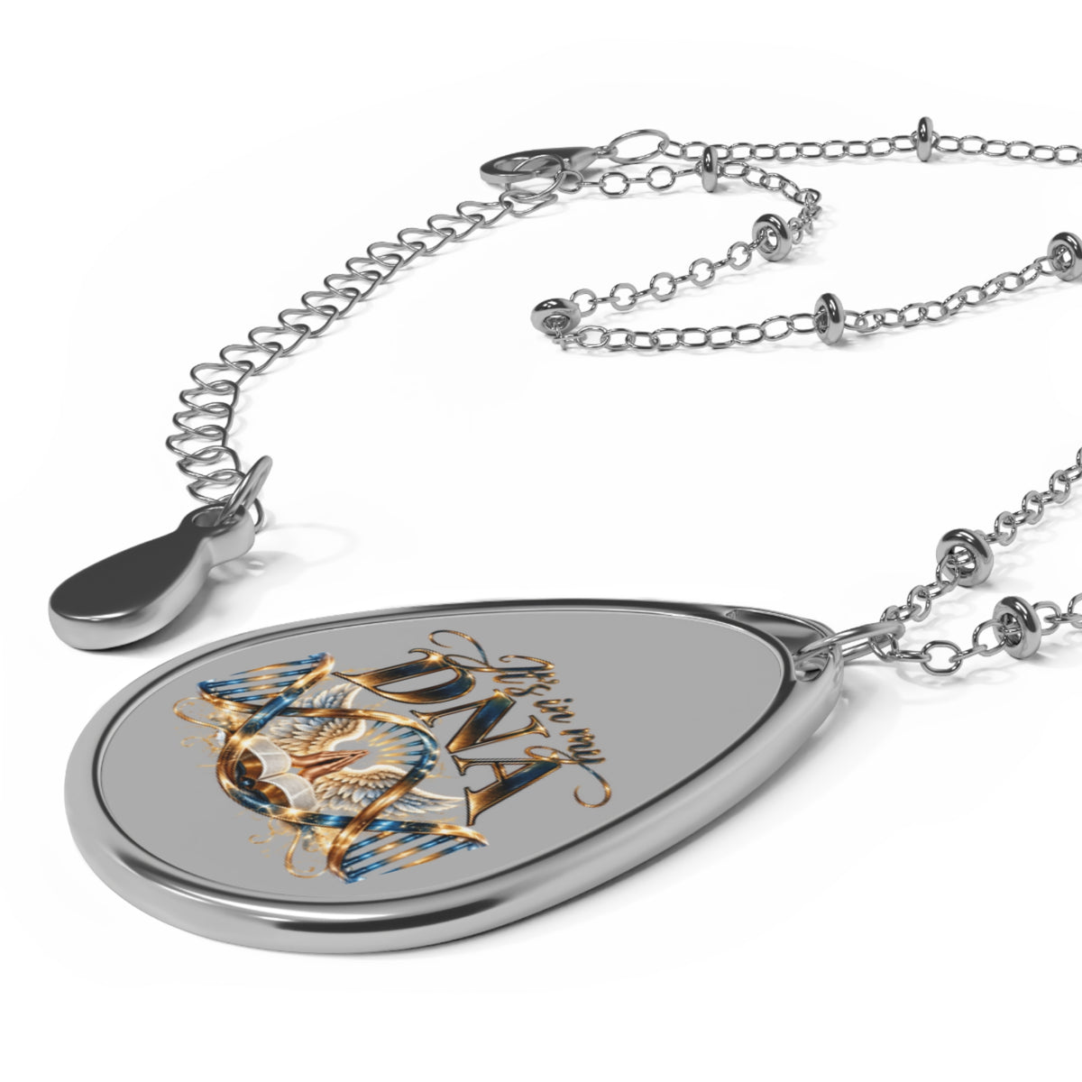 A silver teardrop-shaped pendant necklace featuring a graphic design with the text 'It's in my DNA' in gold and blue, displayed on a clean white background.